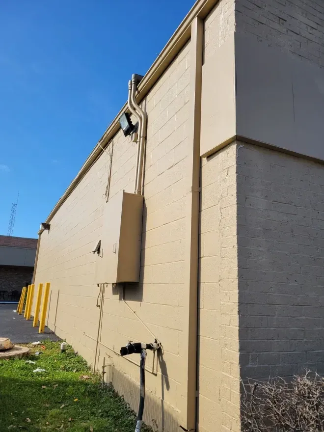 Beige utility building wall with electrical boxes and pipes beside a grassy lot under a blue sky