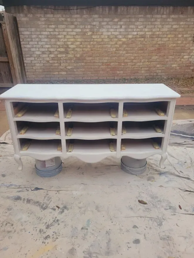 White wooden cabinet with open shelves sitting on buckets in front of a brick wall outdoors