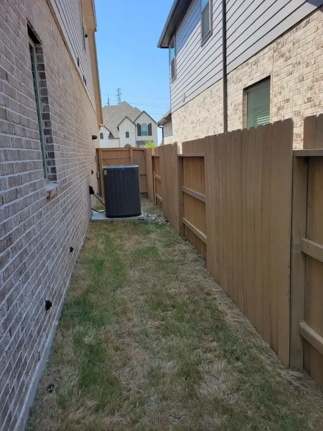 Narrow grassy side yard between two houses, with a wooden fence and outdoor AC unit.