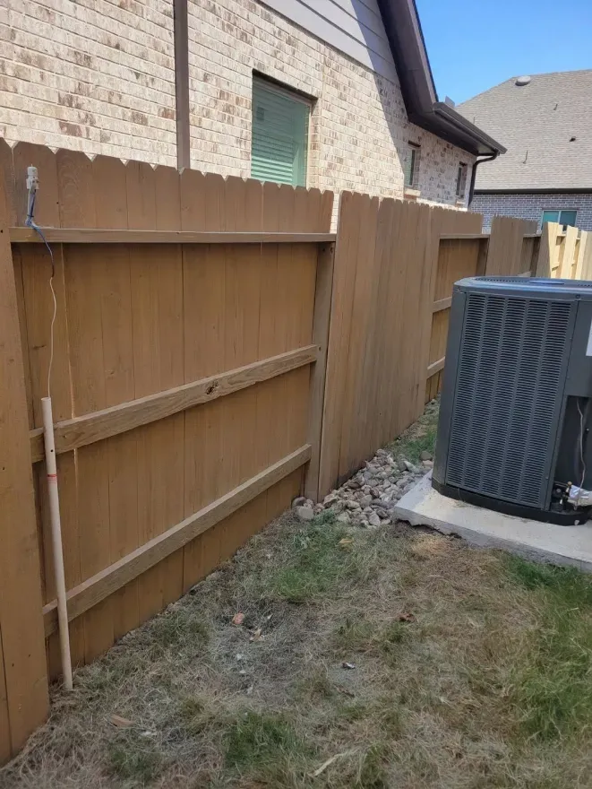 Narrow backyard beside a brown wooden fence and house, with an outdoor AC unit on a concrete pad.