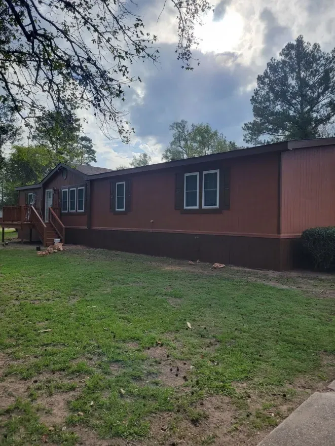 Single-story brown house with a small porch, windows, and a grassy yard under cloudy sky