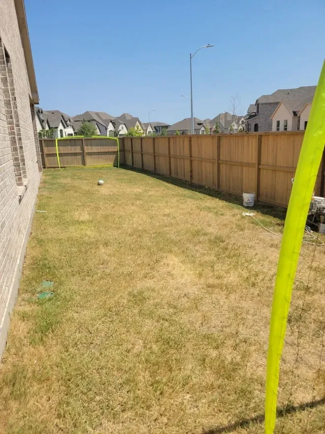 Narrow grassy backyard with a wooden fence, neighboring houses, and a clear blue sky.