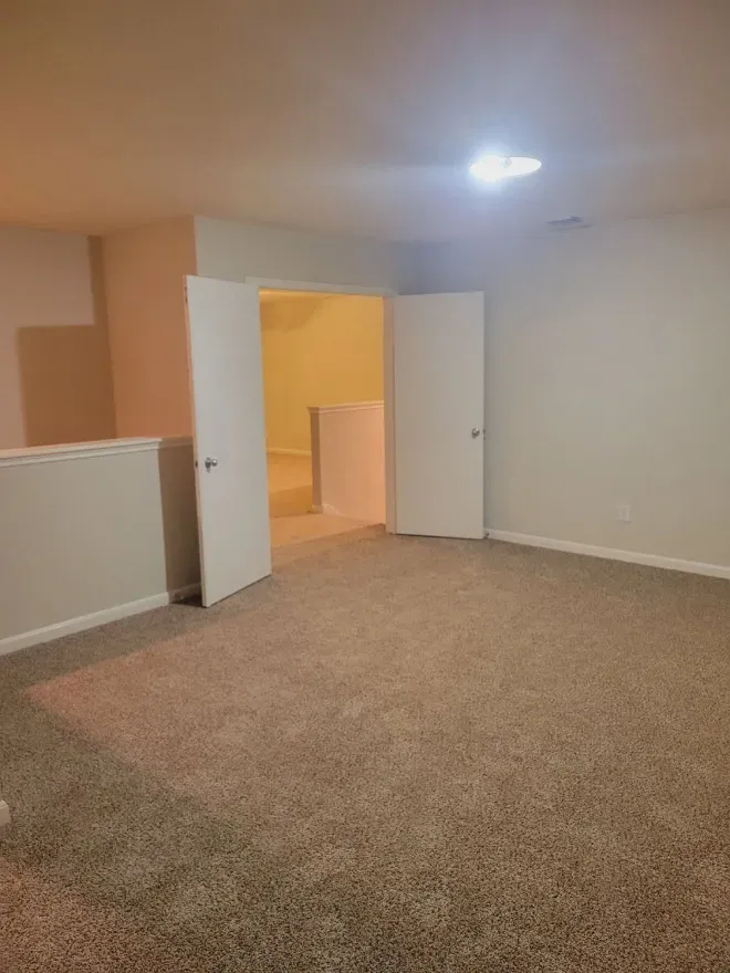 Empty beige carpeted room with open white doors and a ceiling light, leading to a lit hallway.