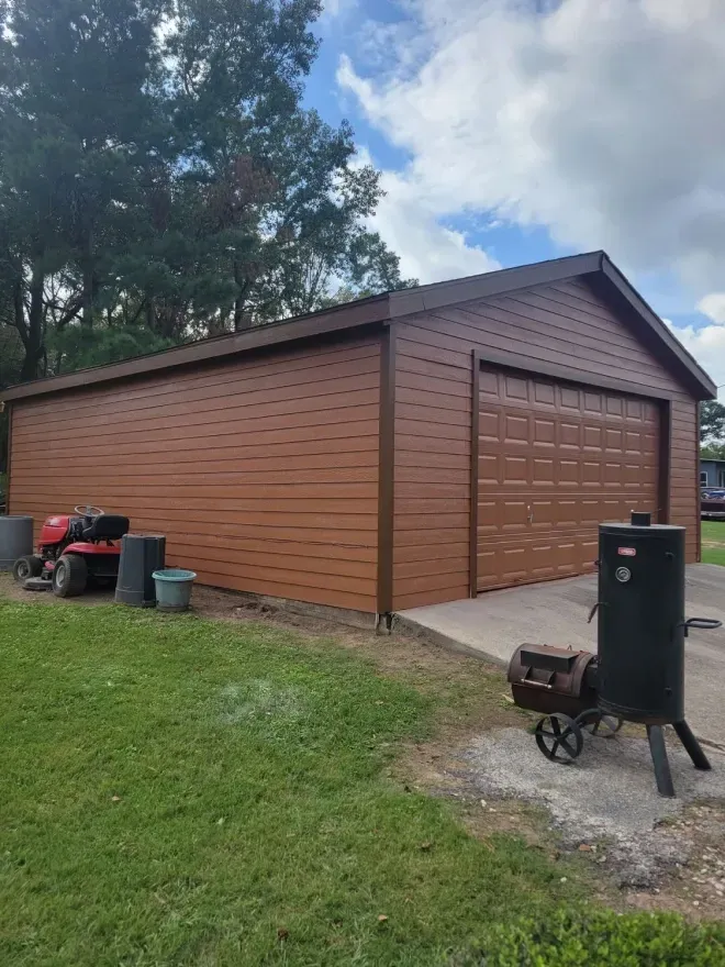 Brown metal garage with large door, trees behind it, and a smoker grill in the foreground on a grassy yard.