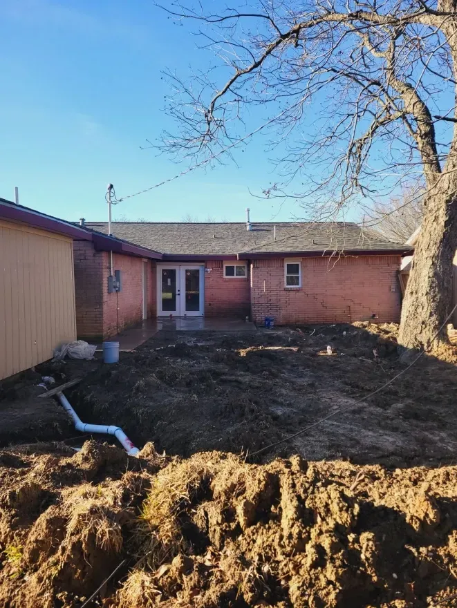 Backyard with muddy dirt pile, white pipe, and a brick house under a clear blue sky