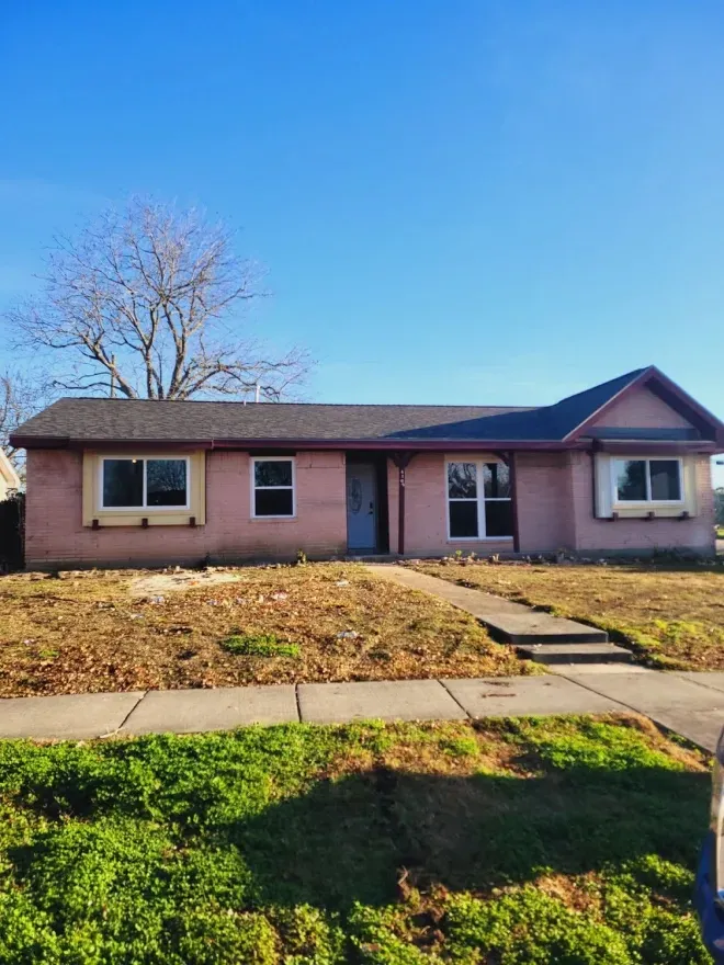 Single-story pink house with a front lawn, sidewalk, and bare tree under a blue sky.