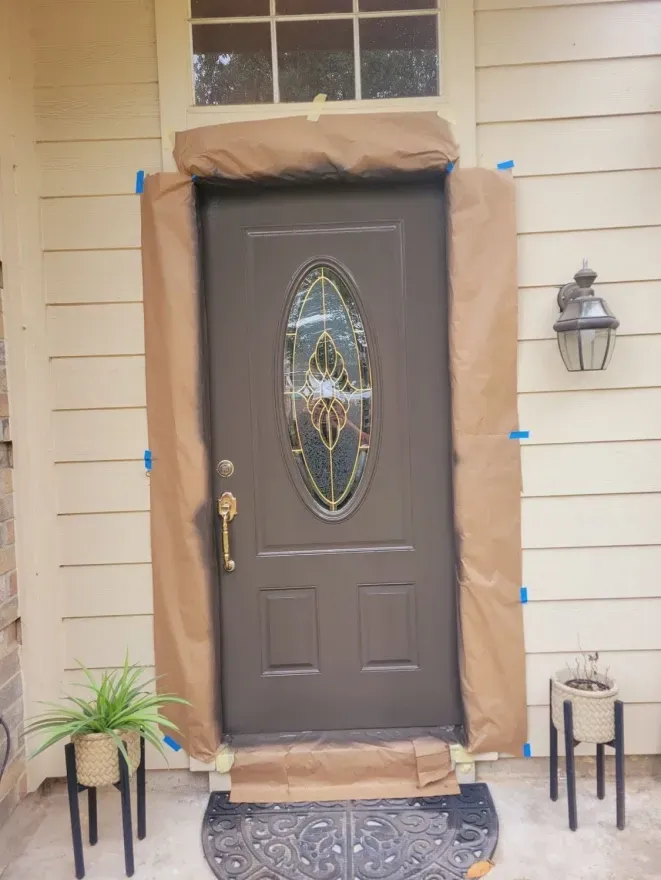 Front door covered in brown protective paper, with glass oval inset, flanked by potted plants and wall lanterns.