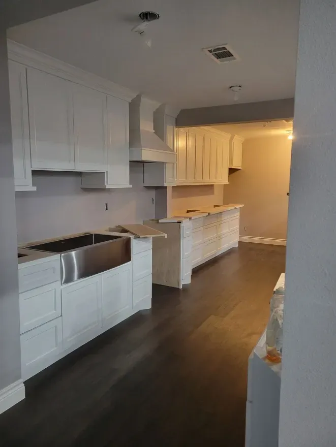 Empty renovated kitchen with white cabinets, dark wood floors, and warm lighting in the adjoining room