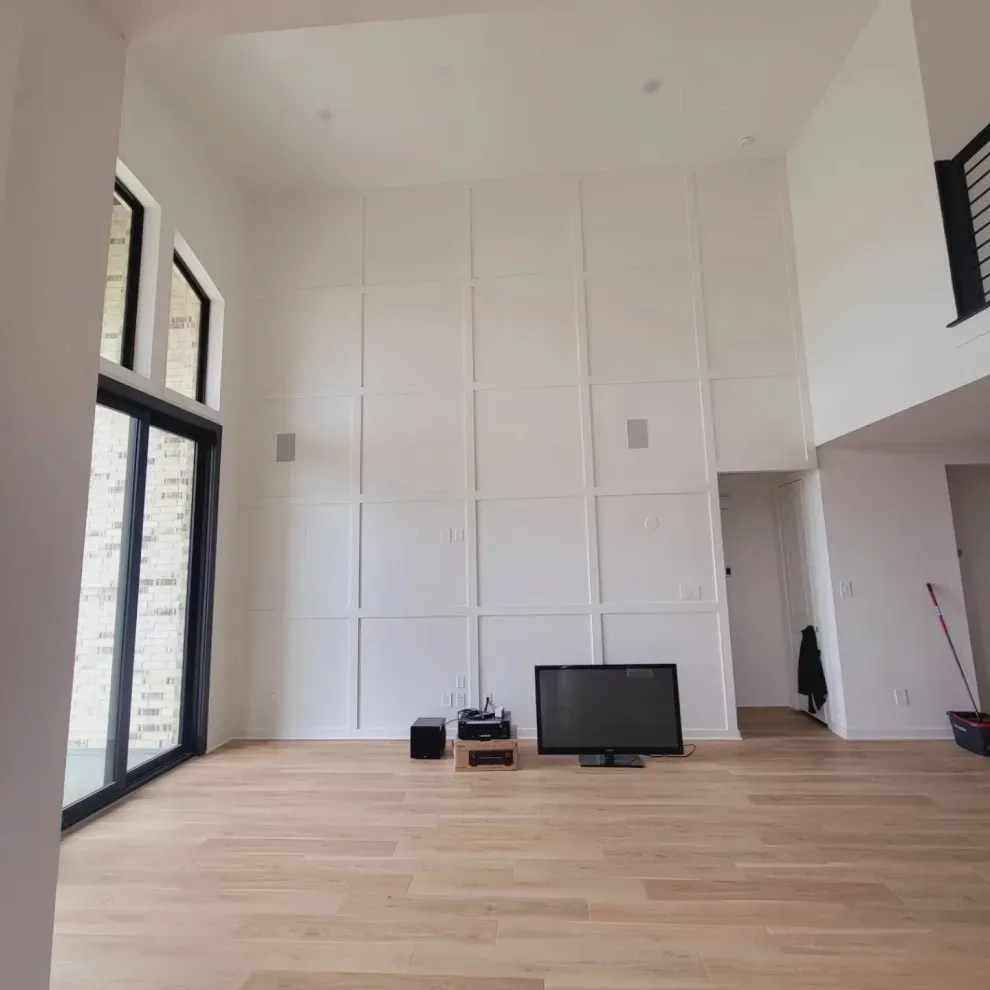 Modern living room with high ceilings, white paneled wall, large windows, and light wood floors