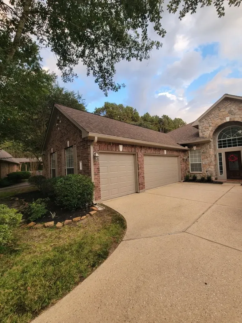 Suburban house with three-car garage, arched stone entry, and curved driveway under trees and cloudy sky