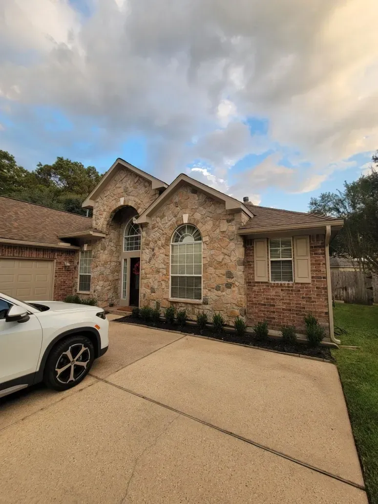 Brick suburban house with arched windows and a driveway, partly showing a white SUV under a cloudy sky