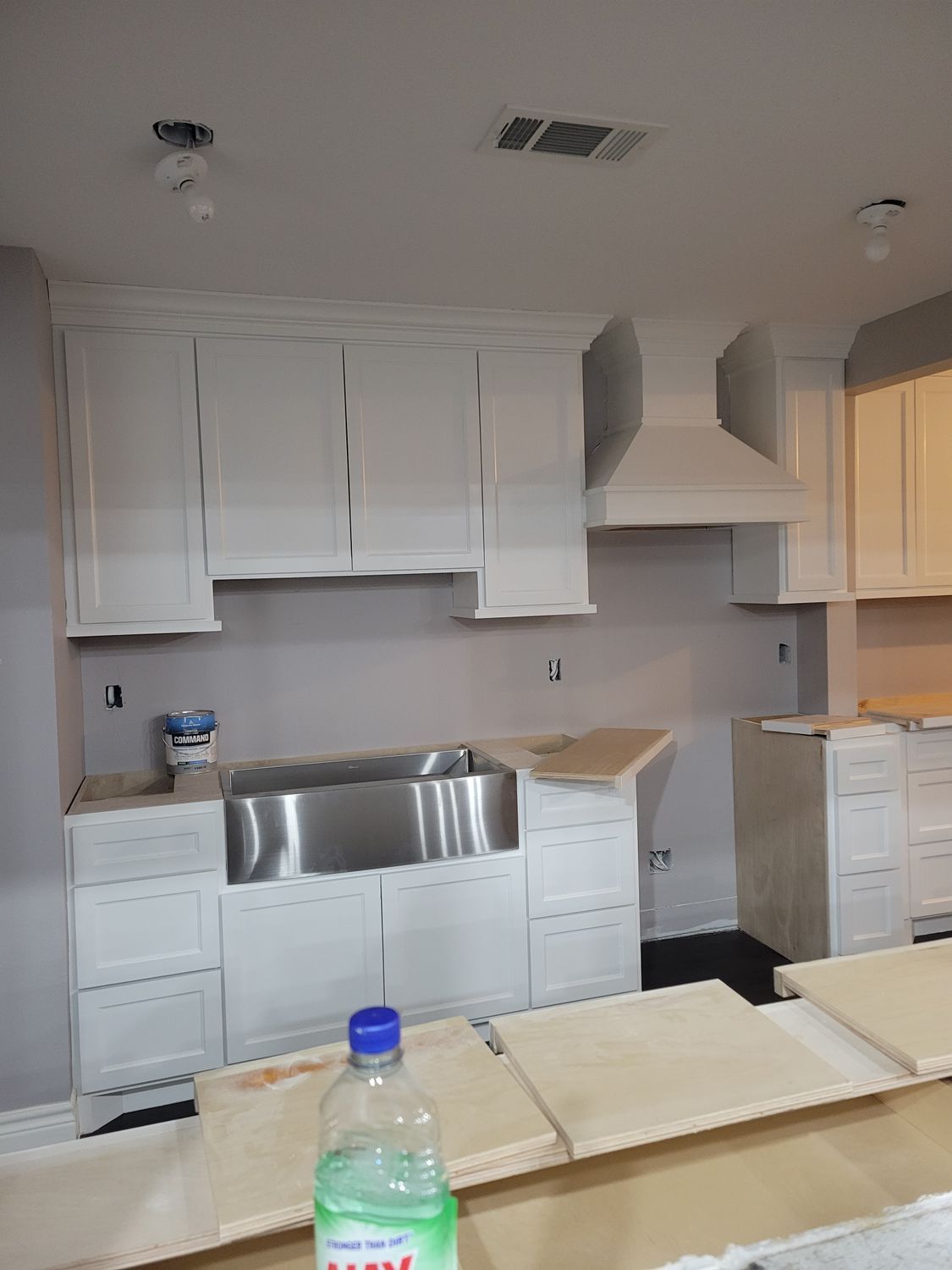 Empty kitchen with white cabinets, stainless sink, and countertop under renovation, with paint supplies on the island.