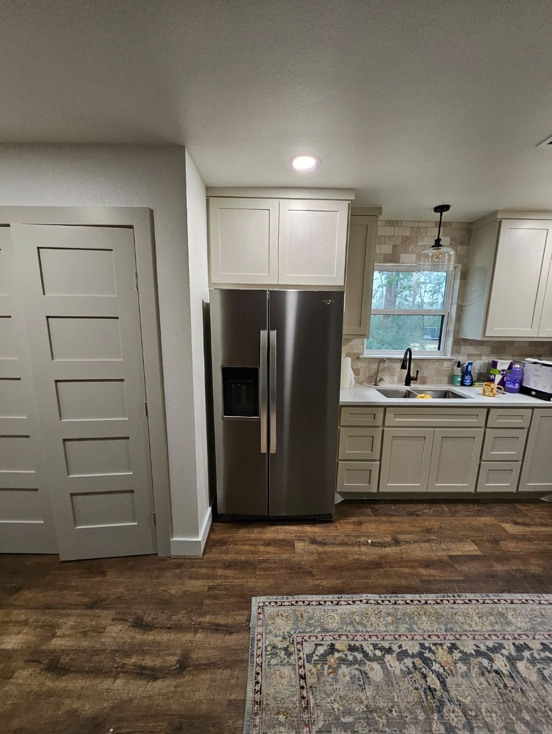 Bright kitchen with stainless refrigerator, white cabinets, wood floor, and a view of the sink and window.
