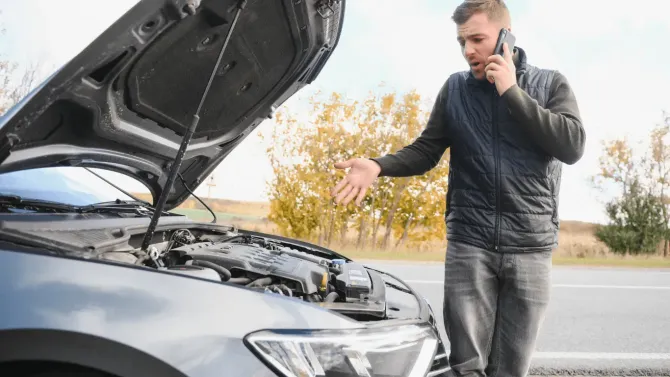 Man on phone beside open car hood, checking engine on roadside.
