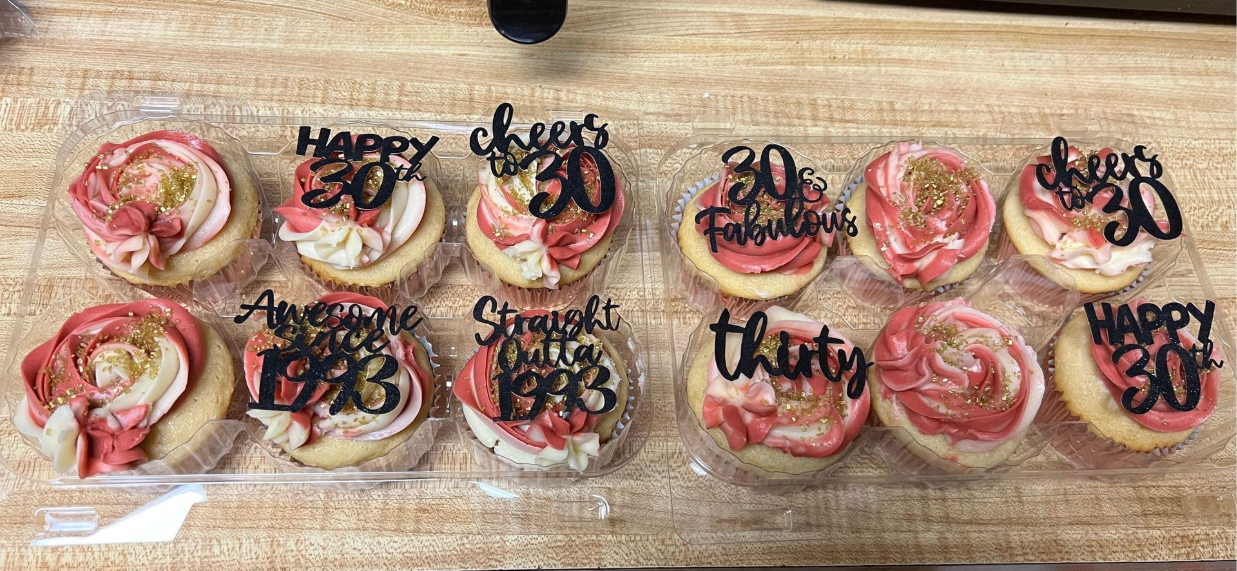 Assorted decorated cupcakes on a wooden tray, with pink frosting and black celebratory lettering.