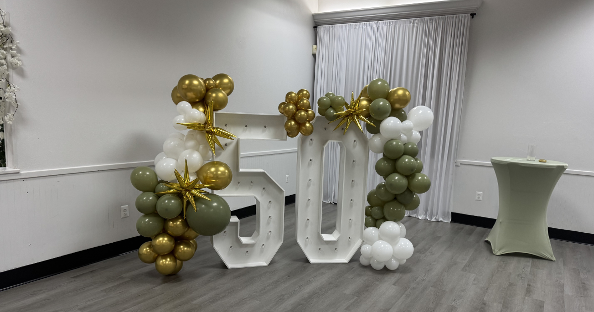 Balloon-decorated white marquee letters in a bright room, with gold, white, and olive-green balloons.