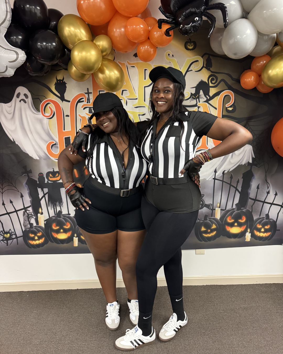 Two people in black-and-white striped outfits posing at a Halloween party backdrop with balloons and pumpkins.