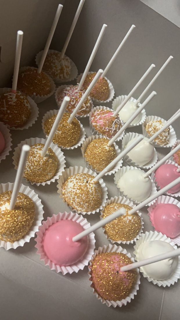 Assorted pink, white, and gold cake pops in white paper cups, arranged in a box.