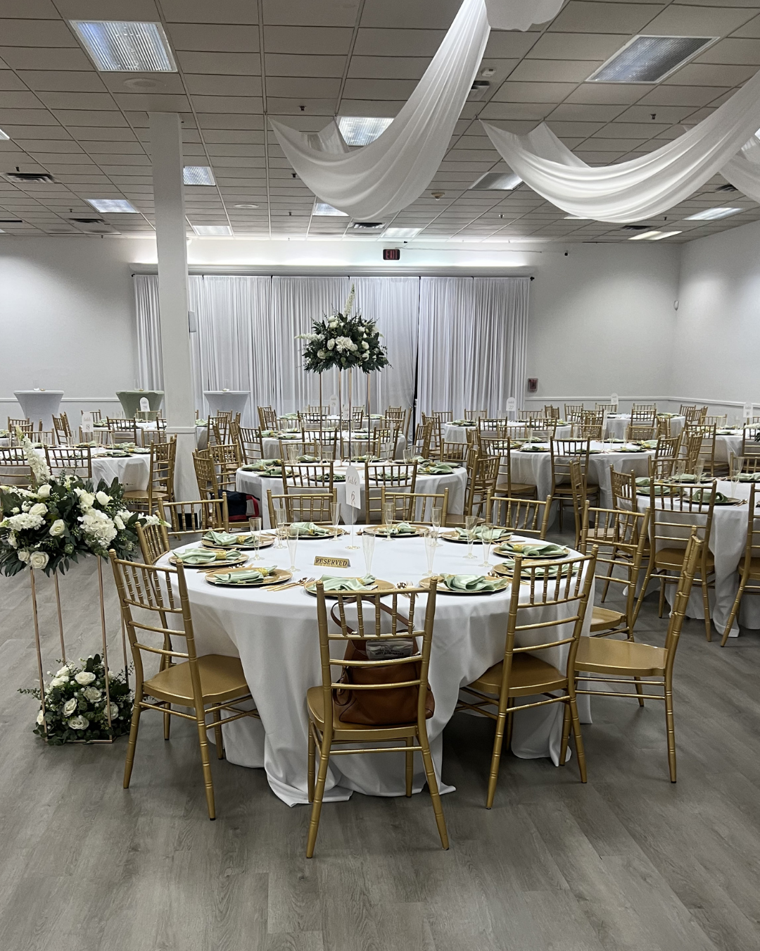 Elegant banquet hall with round tables, gold chairs, white drapes, and floral centerpieces prepared for an event