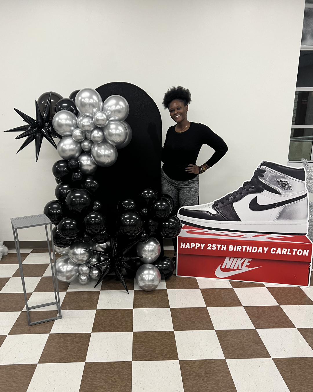 Person posing beside black-and-silver balloon display and oversized Nike shoe box on a checkered floor.