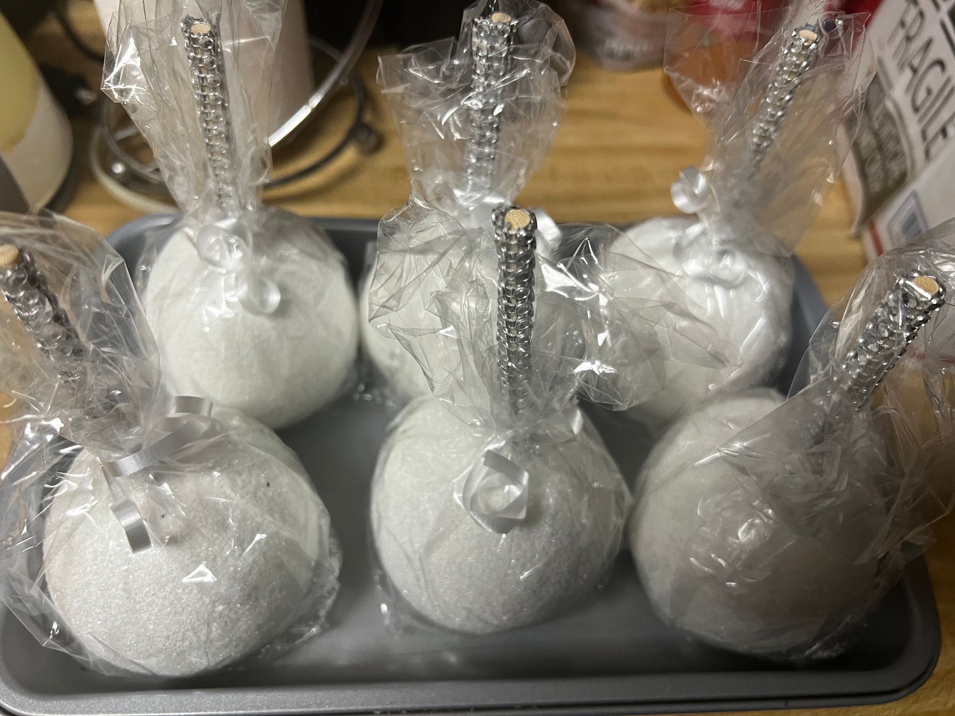 Tray of white spherical treats wrapped in plastic, each with a silver stick, on a countertop