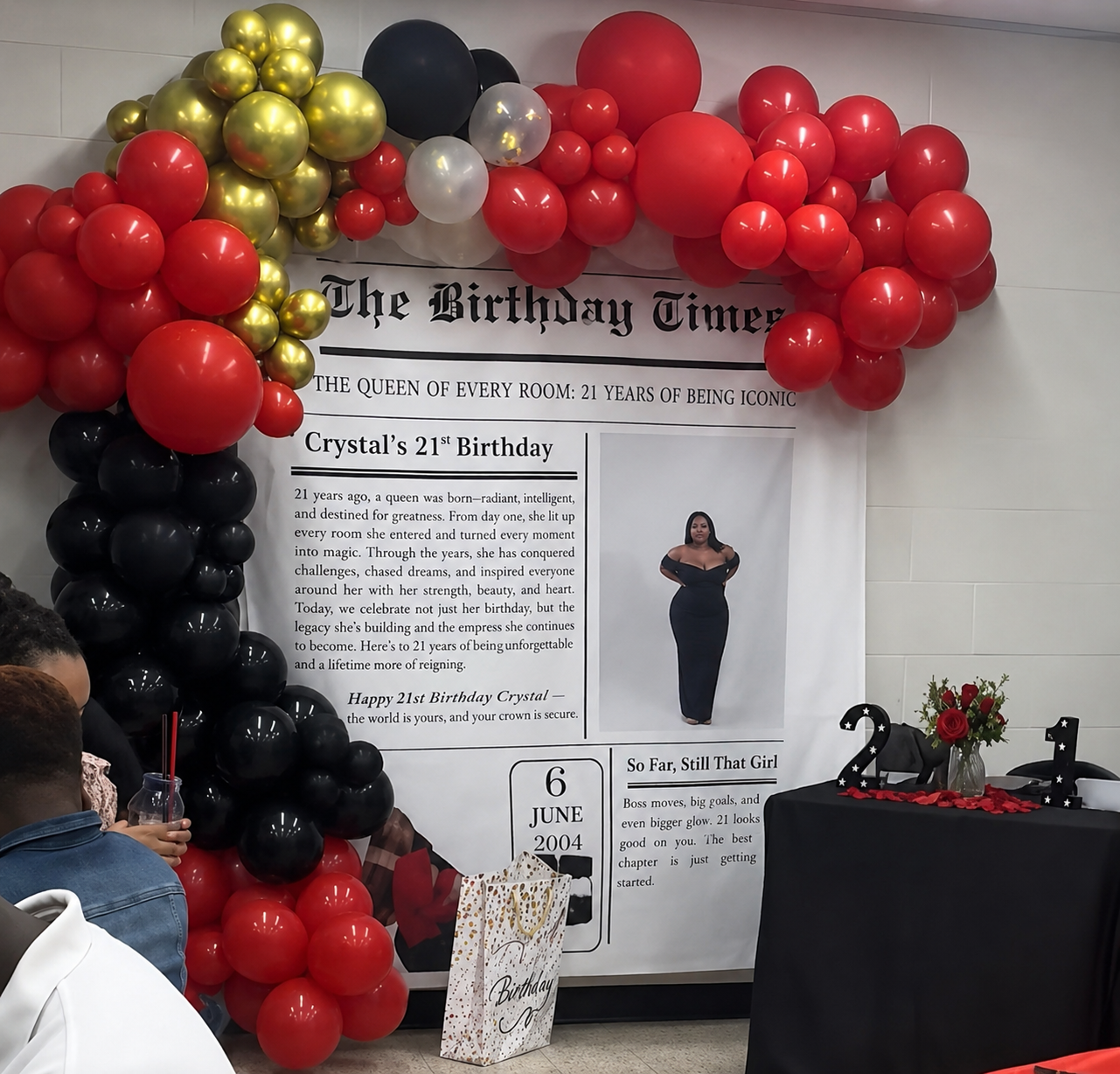 Birthday display with black, red, and gold balloons, a banner, and a tribute poster on a decorated wall