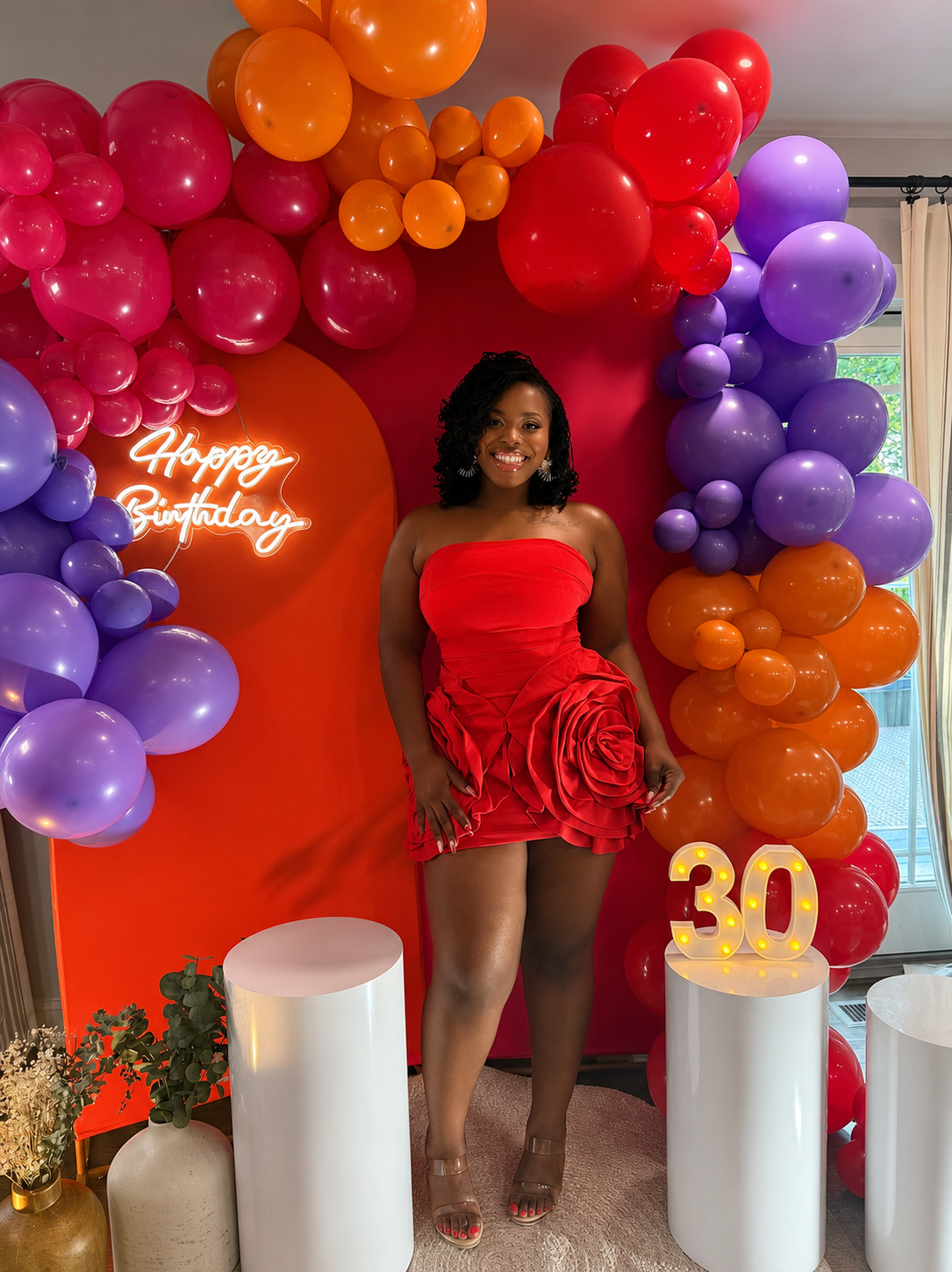 Woman in red dress posing in front of pink, purple, and orange balloon backdrop with “Happy Birthday” and “30” decor