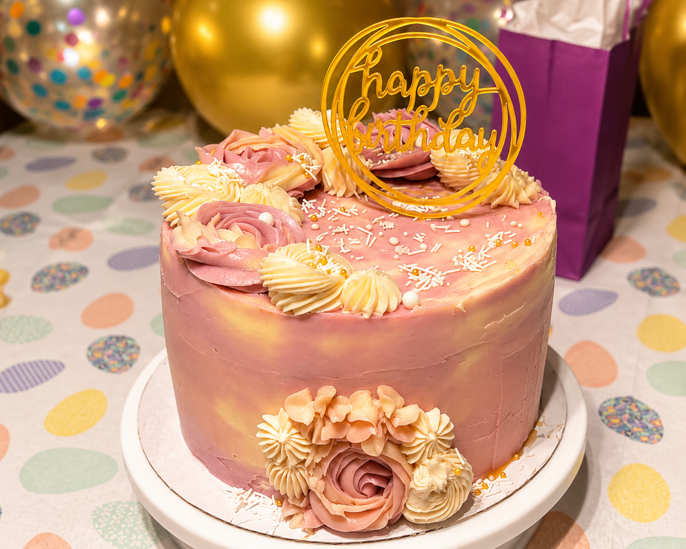 Pink frosted birthday cake with gold floral decorations and topper on a white stand at a party table