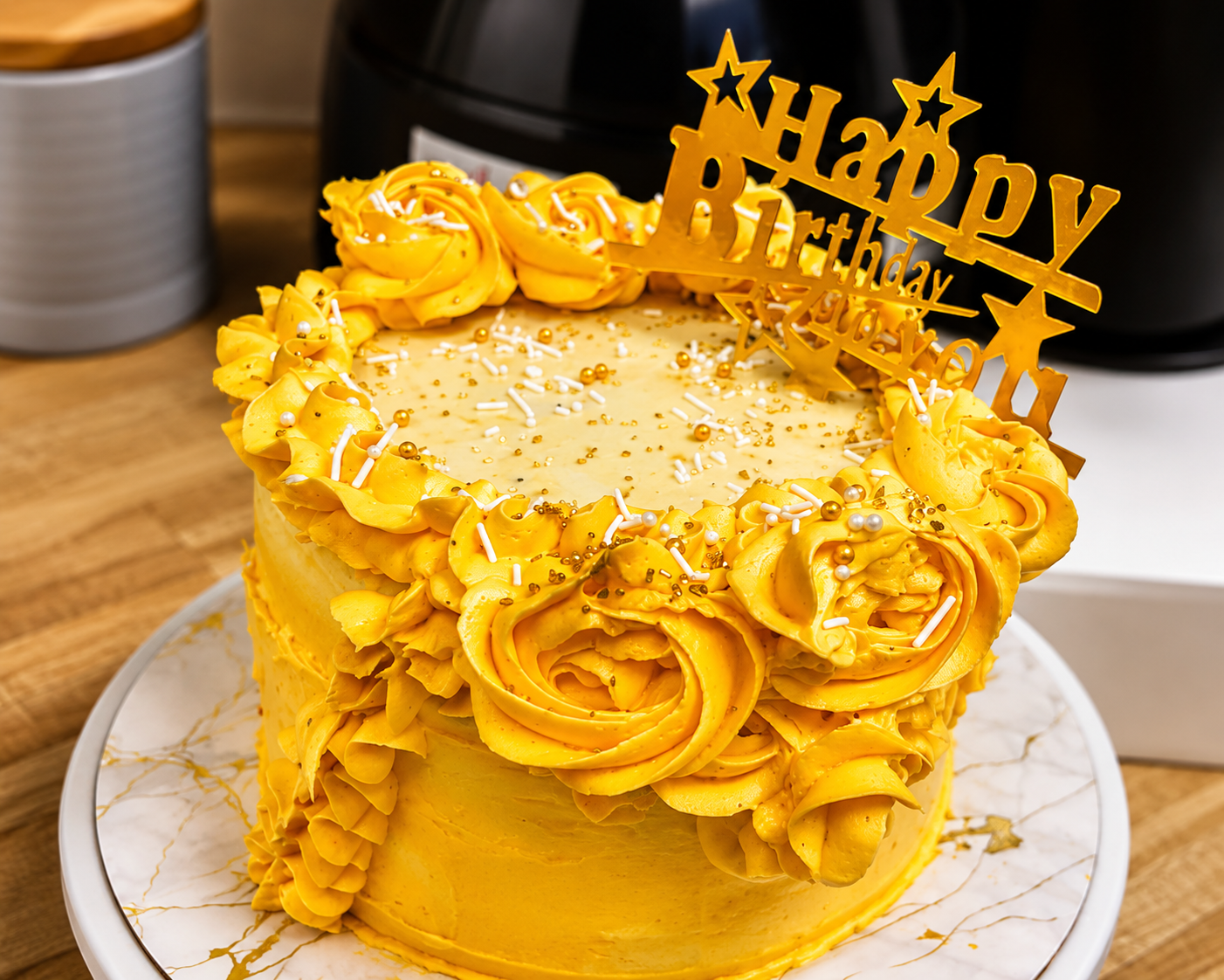 Yellow birthday cake with gold rosettes and “Happy Birthday” topper on a white plate