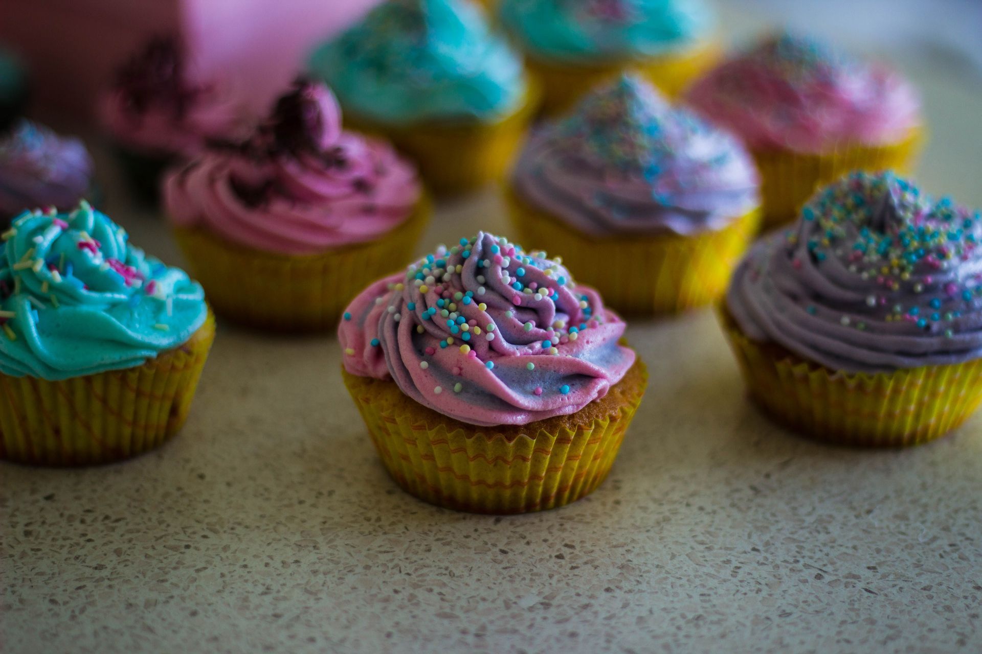 Colorful frosted cupcakes with sprinkles on a countertop, one pink cupcake centered in the foreground