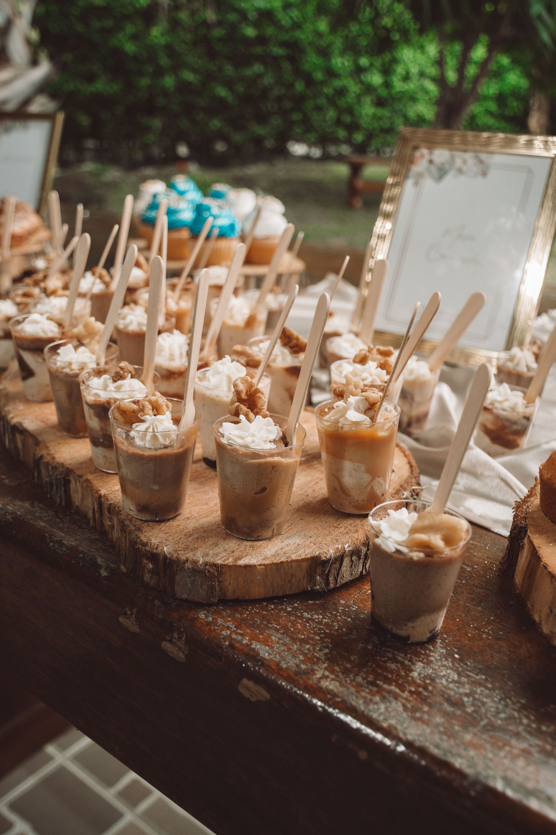 Dessert cups with spoons on a rustic table at an outdoor event, with a gold-framed sign behind them