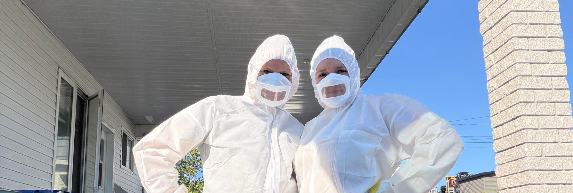 Deux personnes en tenue de protection blanche posent devant une maison.