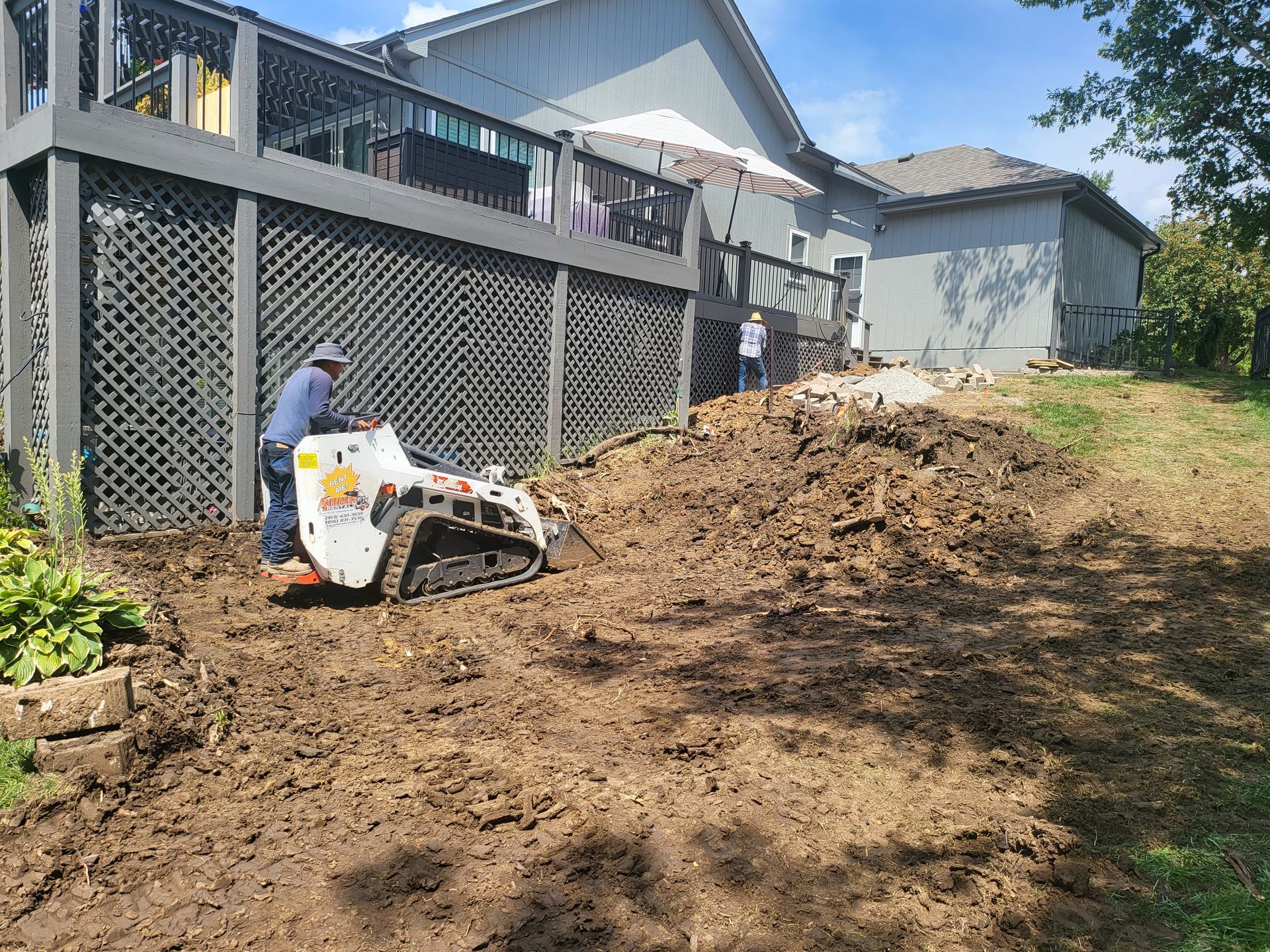 Worker operating a small loader beside a house, moving dirt along a sloped yard
