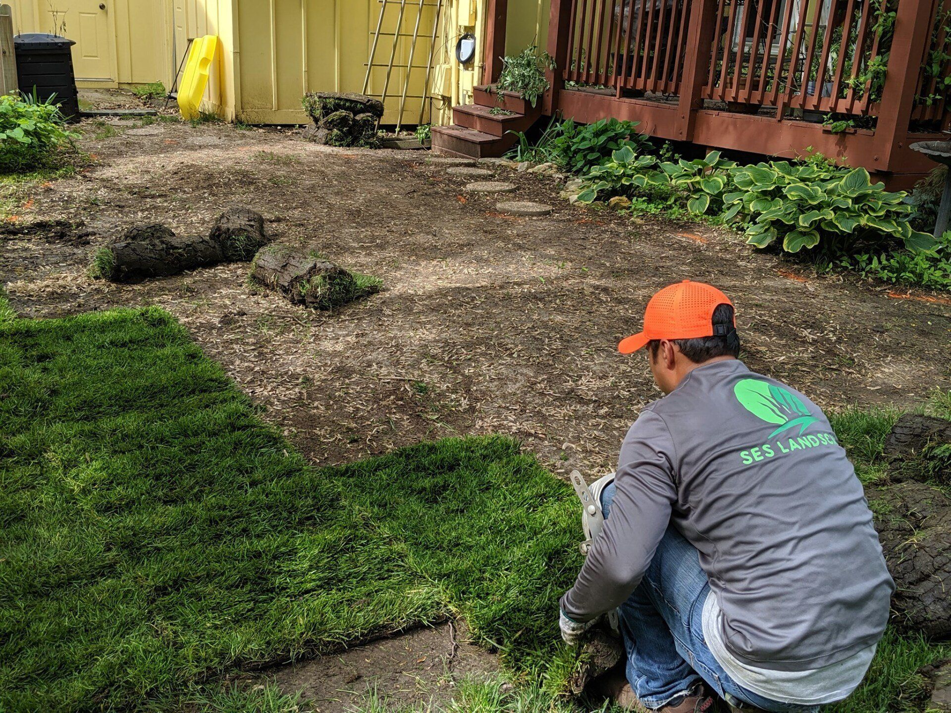 Worker kneeling on a yard, laying sod beside a yellow house and wooden deck.
