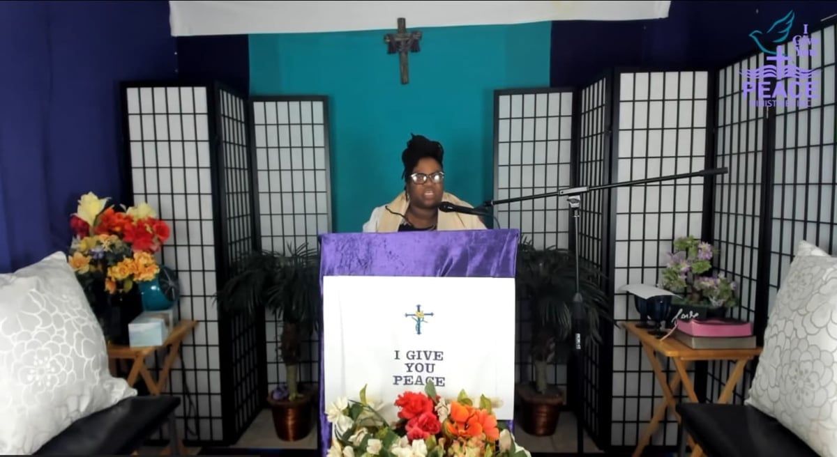 Person speaking at a pulpit in a small church, with floral arrangements and folding screens behind them.