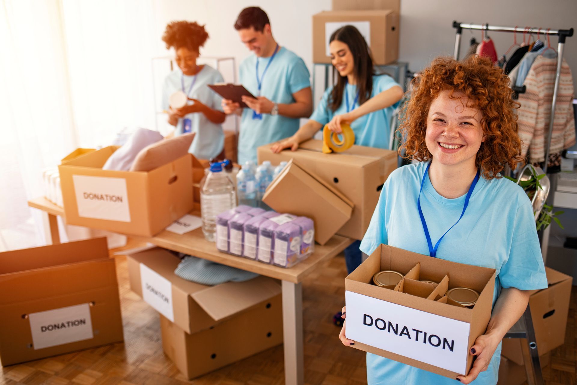 Volunteers packing donation boxes in a bright room, with stacked supplies and smiling people sorting items.