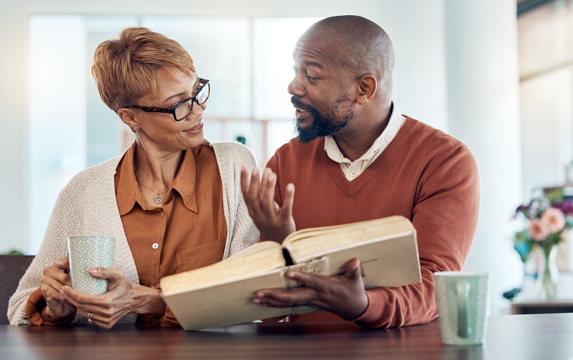 Two people sit at a table, smiling and discussing an open book, with drinks beside them.