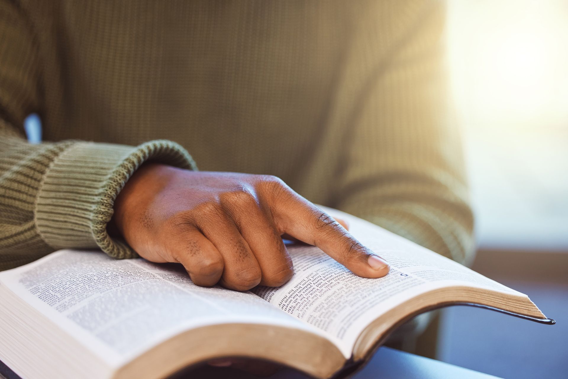 Hand pointing to text in an open book, with a beige sweater sleeve visible.
