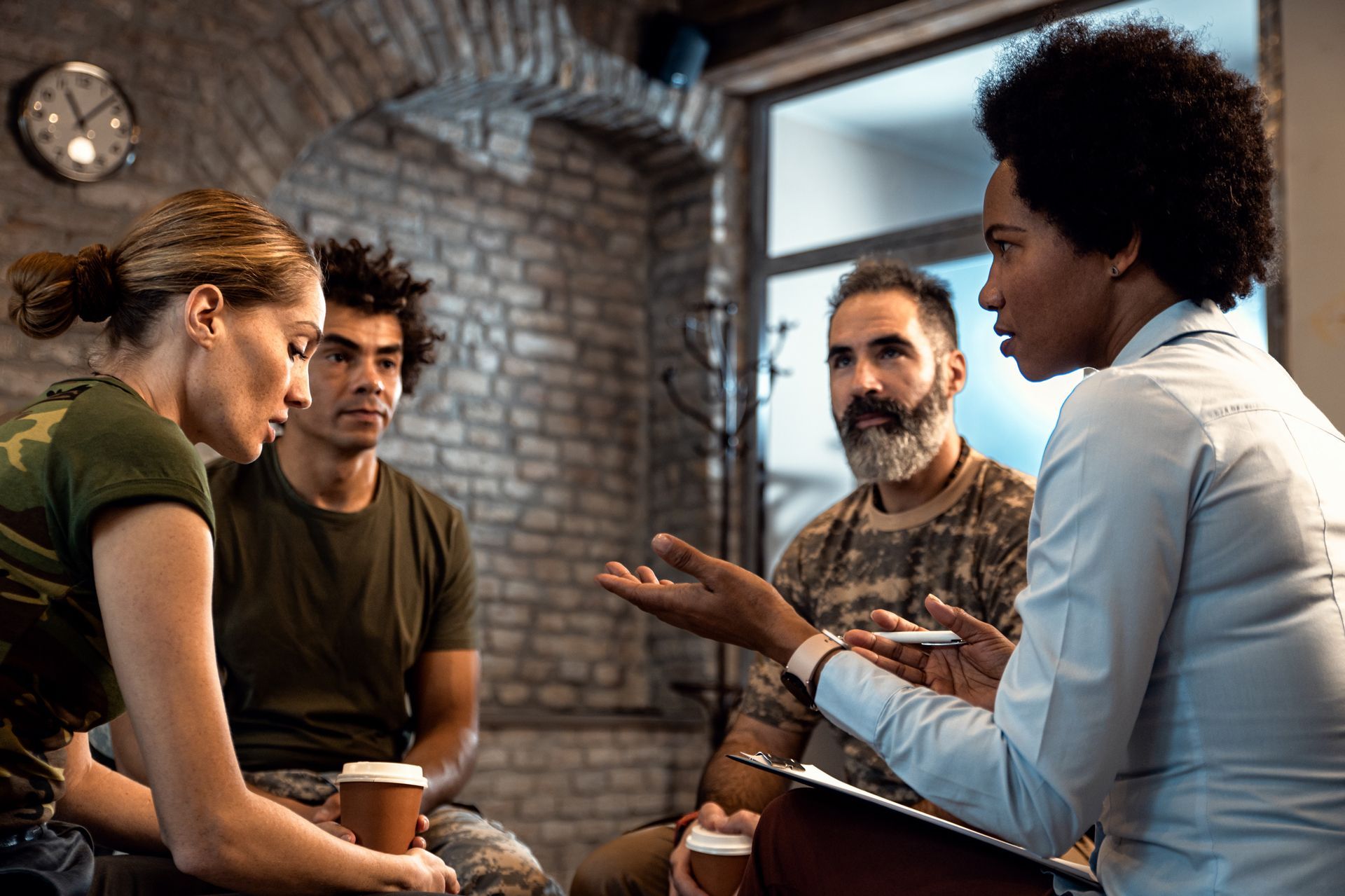 Four colleagues in a meeting, one woman gesturing while seated in a brick-walled room with coffee cups.