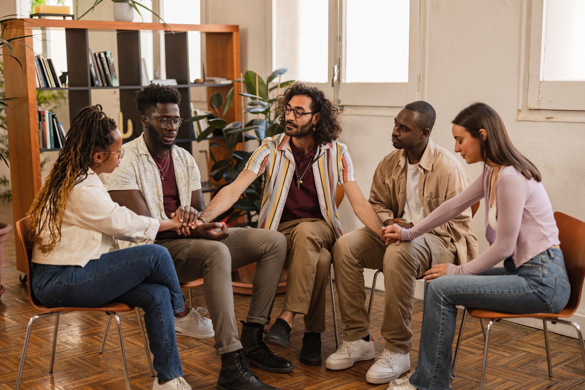 Five people sit in a circle talking in a bright room with shelves and plants.