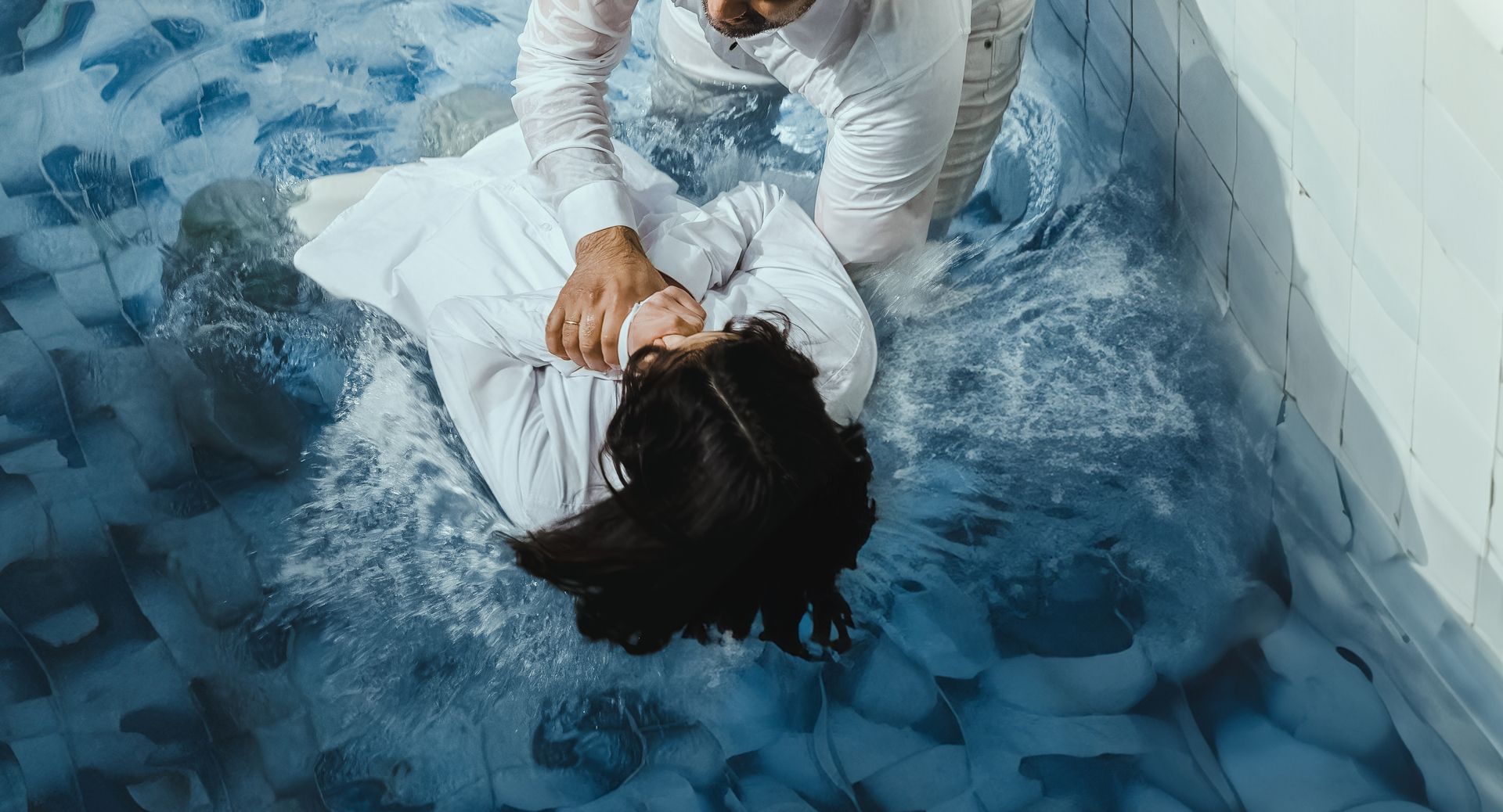 Person in white clothes lies on a blue-tiled floor as another kneels over them, holding their neck.