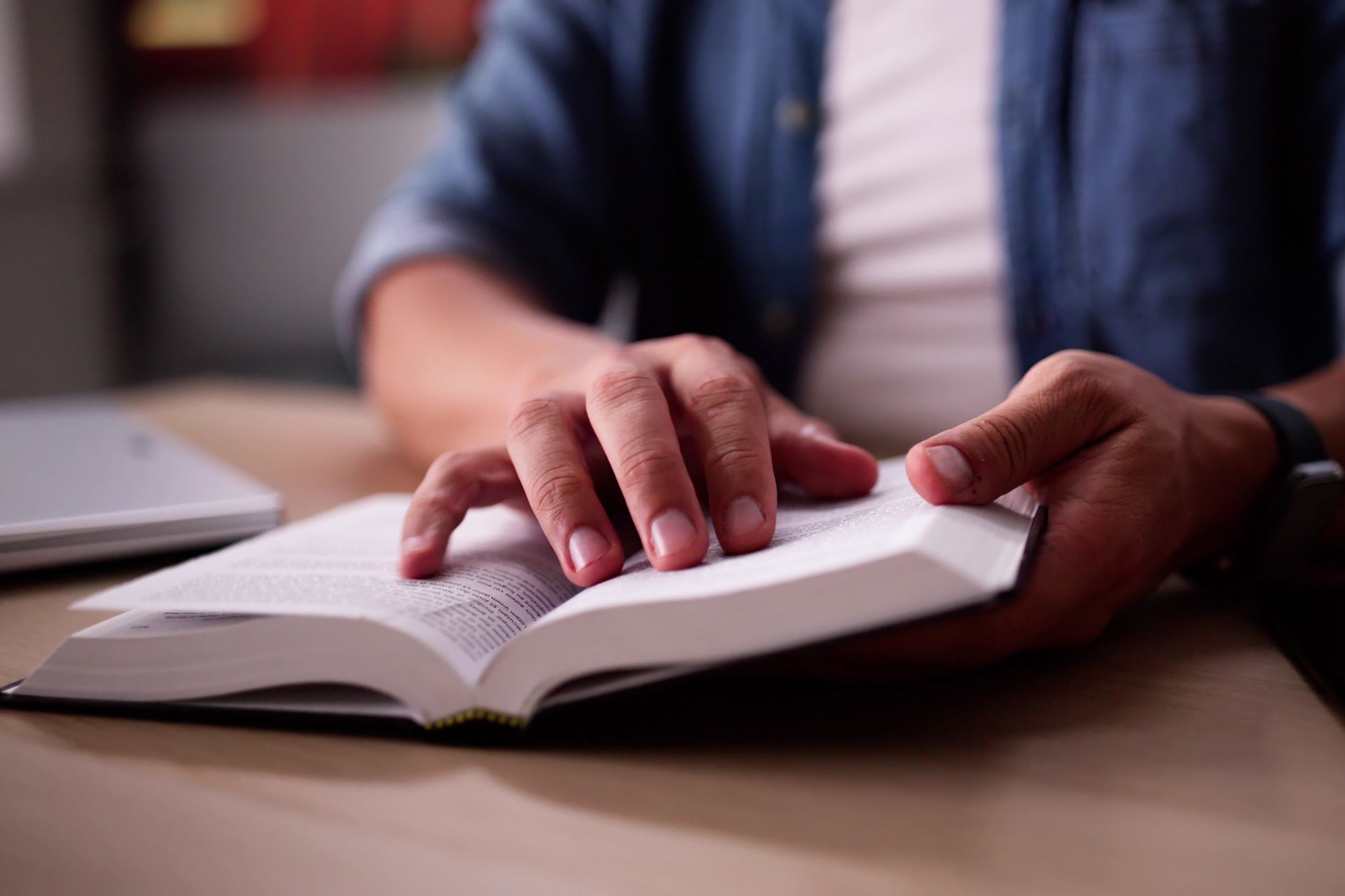 Hands reading an open book at a table, with a blurred person in a blue shirt in the background