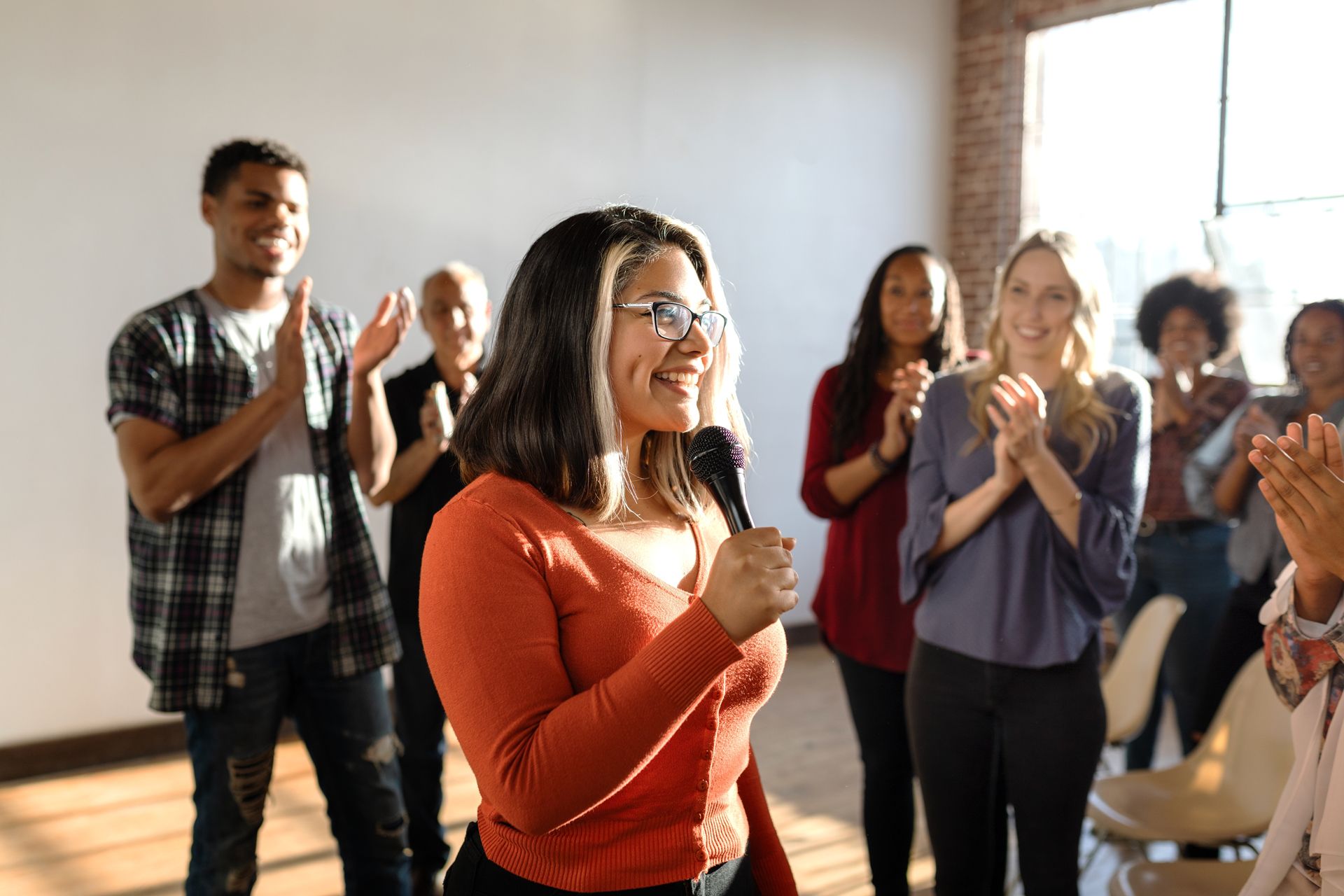 People applauding in a bright room, with a woman in an orange top speaking into a microphone