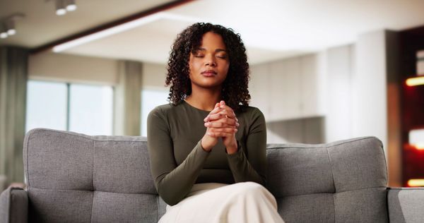 Woman seated on a gray sofa, hands clasped, in a bright living room.
