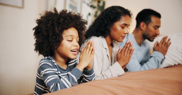 Three people sitting at a table with hands clasped in prayer, smiling in a bright room