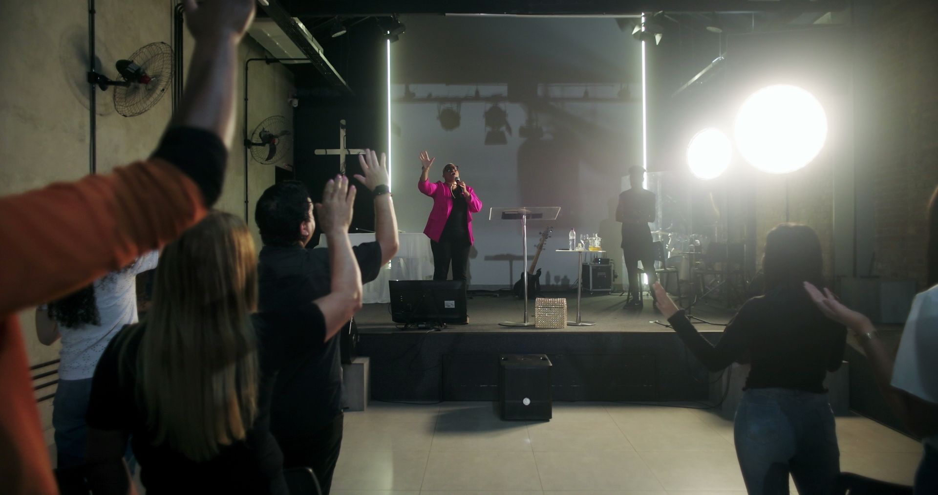 Singer in bright pink jacket performing on a small stage, with audience raising hands in a dim room