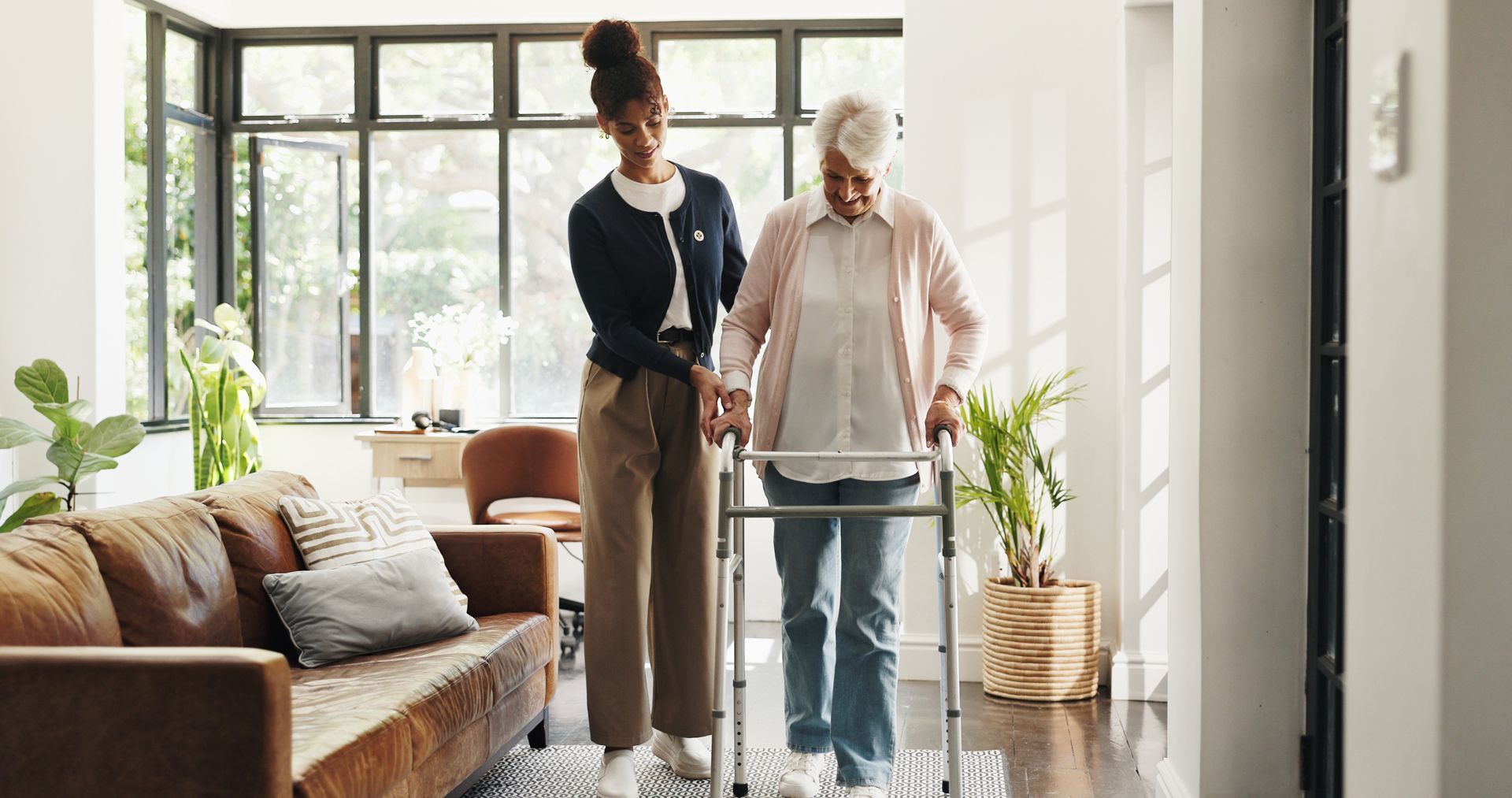 Two people walk with a walker in a bright living room beside a brown sofa and large windows