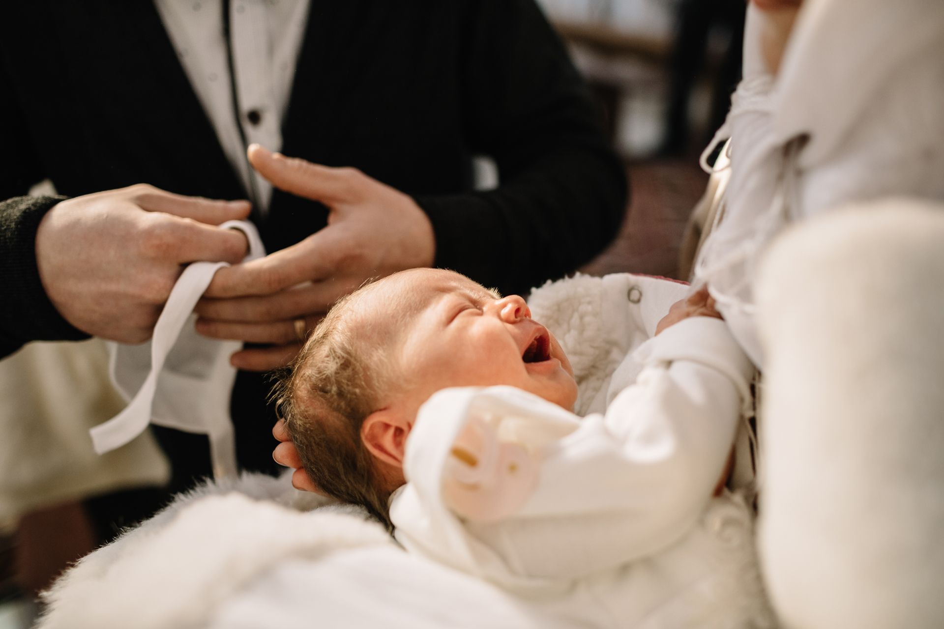 Person fastening a white garment while a baby sleeps nearby on a white cushion