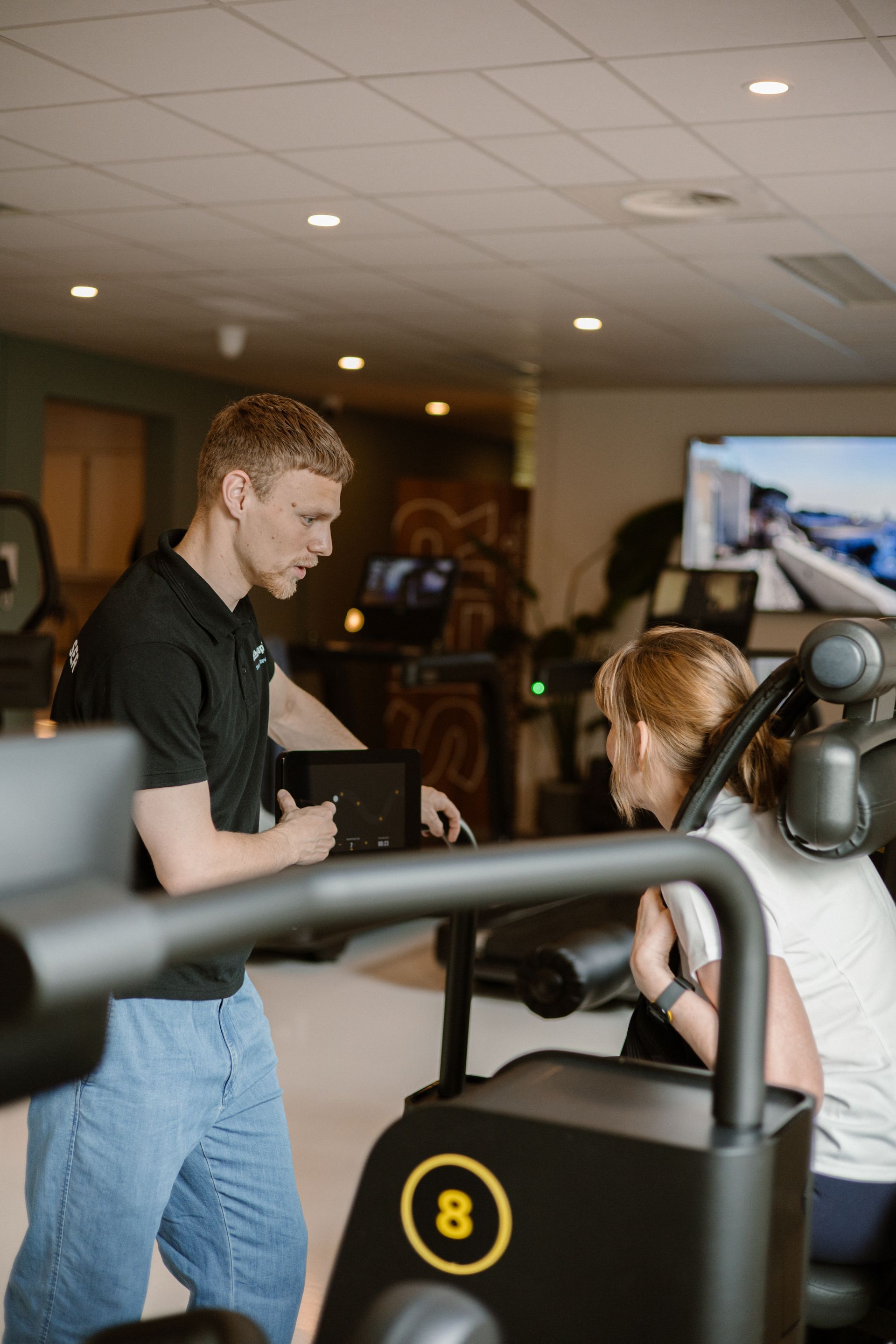 Twee mensen in een binnengymzaal, de een gebogen over een fitnessapparaat terwijl de ander er vlakbij staat.