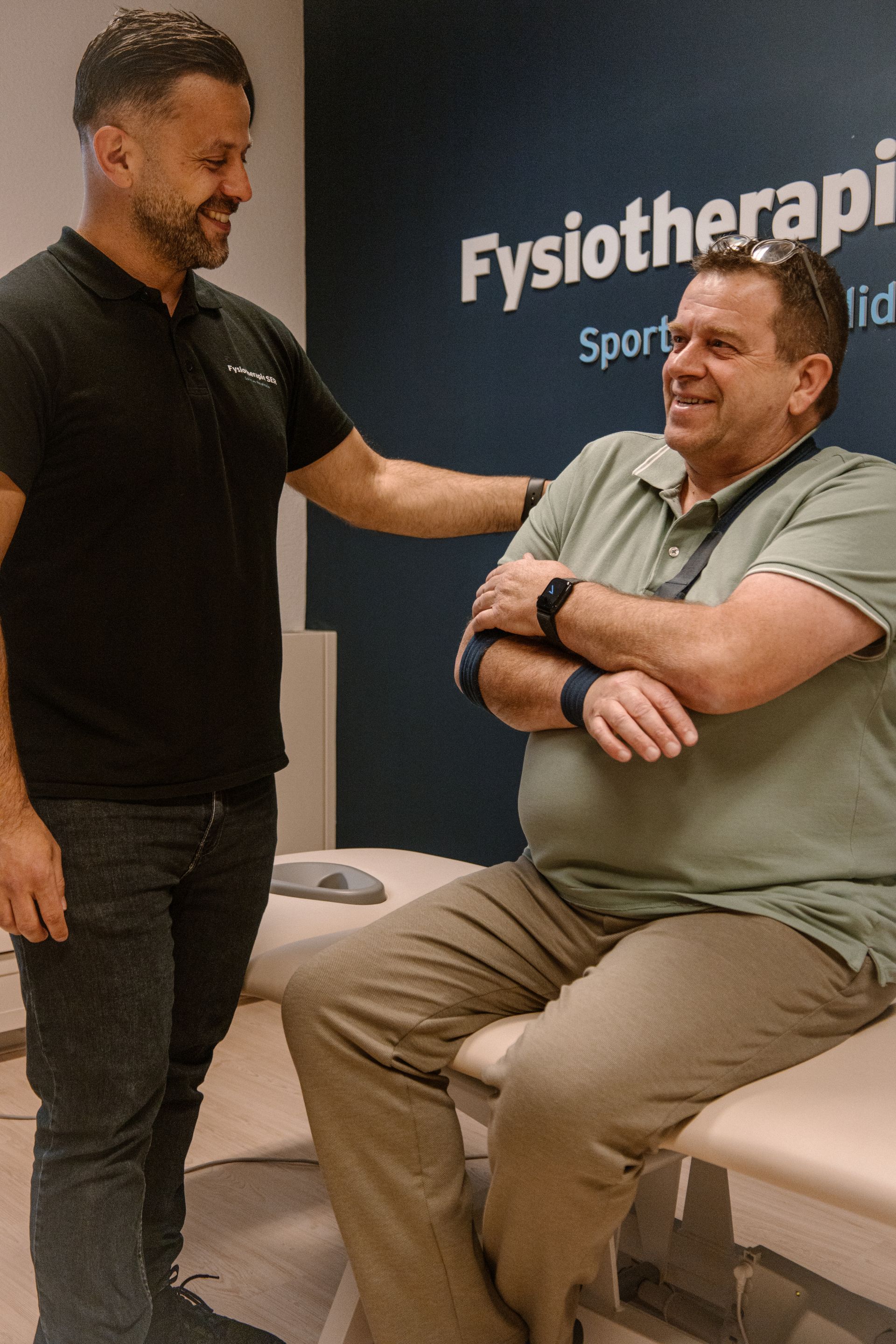 Twee mannen in een fysiotherapiekliniek, waarvan er één staat en glimlacht terwijl hij praat met een zittende patiënt met gekruiste armen.